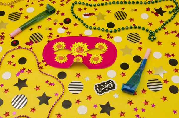 Top view decoration for party. Pink eye mask, plastic necklaces, party blowers and circles and stars confetti on yellow background - Photo, Image