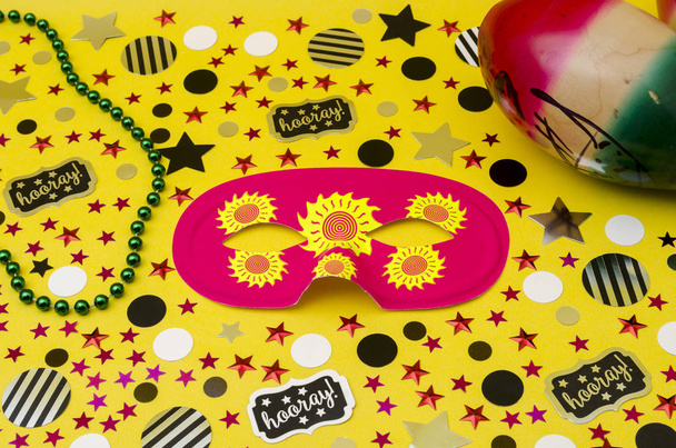 Top view decoration for party. Pink eye mask, plastic necklaces, party blowers and circles and stars confetti on yellow background - Photo, Image