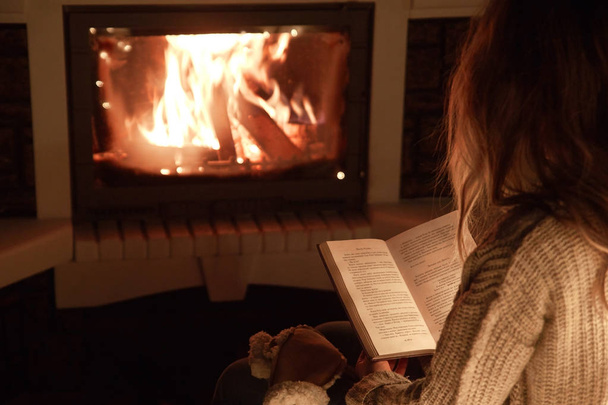 young girl reads a book in front of a fireplace in the evening at home - Photo, Image