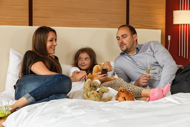 Young happy smiling Caucasian family of father, mother and daughter lying together on bed in hotel bedroom while having vacation at resort - 写真・画像