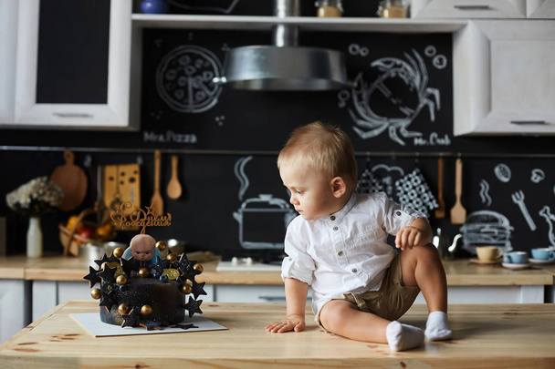 Cute little stylish baby boy in white shirt and shorts sits on the table, looking at a birthday cake and posing at the kitchen interior - Photo, Image