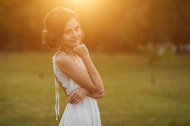 Beautiful and young brunette model girl with gentle makeup, in short white dress stands and posing at green park in early evening at the sunset - Fotografie, Obrázek