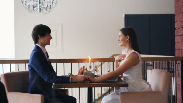 Side άποψη του ευτυχισμένο ζευγάρι στο εστιατόριο κατά τη διάρκεια της πρότασης γάμου, ο τύπος μιλά και βάζοντας δαχτυλίδι αρραβώνων στο δάχτυλο των κοριτσιών, η νεαρή γυναίκα γελάει και λέει Ναι. - Πλάνα, βίντεο