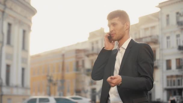 Zakenman op telefoon te praten op het stadsplein met zon flare bij zonsondergang. Jonge man in een zwart suite praten op de mobiele telefoon met handgebaar in de straat warme ochtend zonlicht - Video