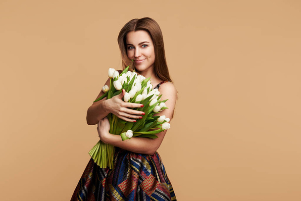 Wow effect. Wonderful flowers on Womens Day. Tender smiling brunet woman poses with white tulips, looks happily, isolated over beige studio background wall. Space for text  - 写真・画像