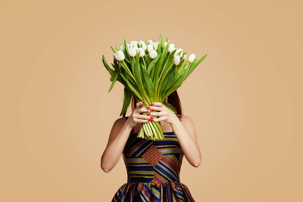 Wow effect. Wonderful flowers on Womens Day. Tender smiling brunet woman poses with white tulips, looks happily, isolated over beige studio background wall. Space for text  - Φωτογραφία, εικόνα