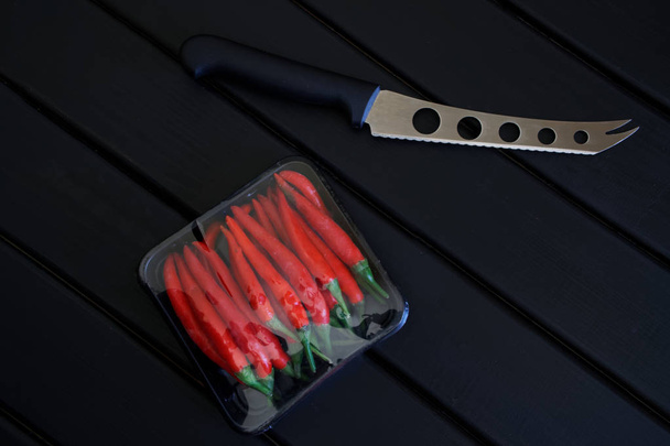 Coltello per formaggi e verdure accanto alla confezione di baccelli di peperoncino rosso si trovano su una superficie di legno nero
.  - Foto, immagini