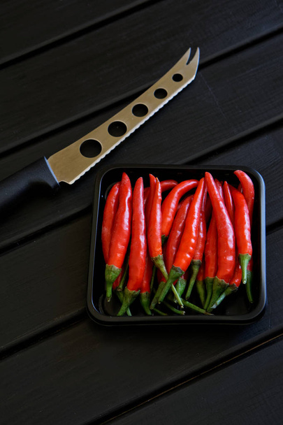 Коробка с красными стручками чили и нож для сыра и овощей лежат на черной деревянной поверхности. Daylight
.  - Фото, изображение