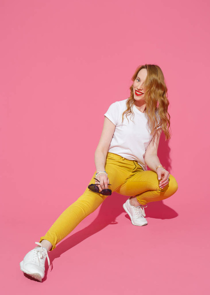 Cheerful blonde woman in trendy yellow jeans and white T-shirt posing on pink background - Photo, image