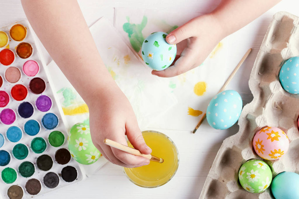 Kid hand painted Easter eggs, paints and brushes on a white table. Preparation for the holiday. Girls hands draw a pattern. - Photo, Image
