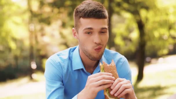 Knappe jonge zakenman eten erg lekker broodje in een zonnig park. Jonge man in een blauw shirt eten van de lunch nevenactiviteiten. Portret shot - Video