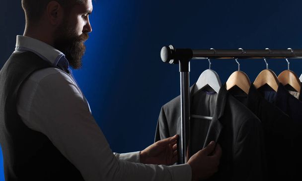 Mens Clothing, shopping in boutiques. Tailor, tailoring. Stylish mens suit. Mens suit, tailor in his workshop. Handsome bearded fashion man in classical costume suit. Male suits hanging in a row - Photo, Image