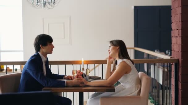Boldog fiatal lány és a fickó beszél tartja kezét ül az asztalnál virágokkal, gyertyákkal és pezsgőt szép étteremben. Ismerkedés és beszélgetési koncepció. - Felvétel, videó