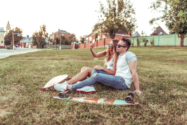 Young couple, selfie photo on phone, video call social networks, online internet application, longboards skate. Happy smiling, concept sporty vacation young family weekends, trendy hipster on grass. - Zdjęcie, obraz
