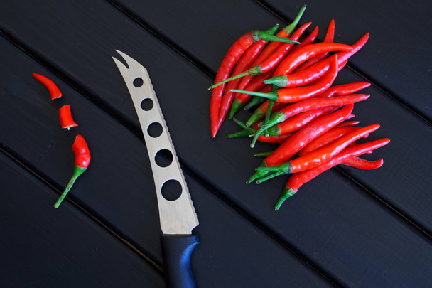 Σε φέτες και το συνολικά κόκκινες πιπεριές τσίλι δίπλα ένα μαχαίρι λαχανικών και τυριών που βρίσκονται σε μια μαύρη ξύλινη επιφάνεια.  - Φωτογραφία, εικόνα