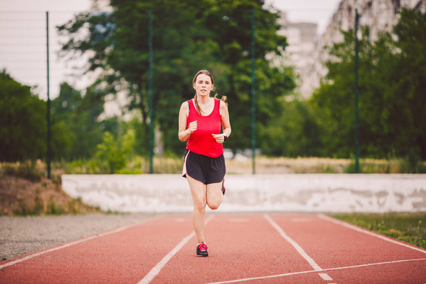 Bella giovane atleta donna caucasica con grandi seni in T-shirt rossa e pantaloncini corti da jogging, in esecuzione nello stadio con rivestimento in gomma rossa
 - Foto, immagini