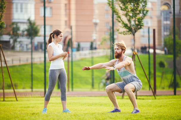 Team work in sports. A female trainer is training a young handsome man with a beard in a park on green grass, a lawn at sunset. A girl holds a phone in her hand and uses a stopwatch - 写真・画像