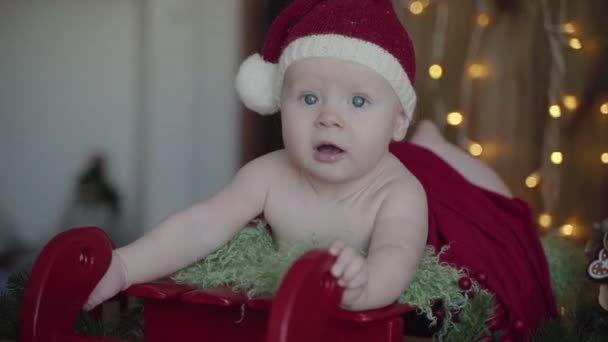 A little baby lying on the Santa Claus sleigh in a Christmas hat and suit, New Year photoshoot - Filmagem, Vídeo