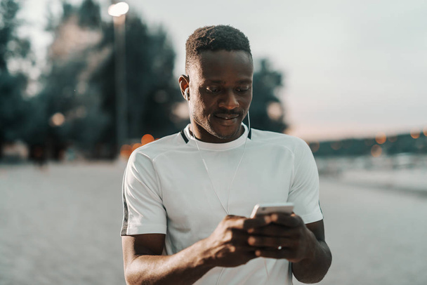 Smiling fit african man in white t-shirt putting earphones in ears and holding smart phone. - Photo, Image