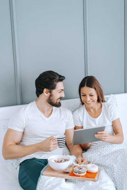 Jovem feliz casal caucasiano apaixonado tomar café da manhã na cama e assistir a vídeos em tablet
. - Foto, Imagem