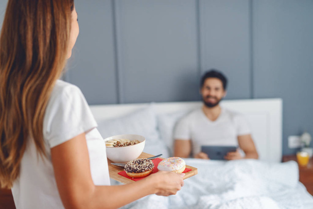 Atrakcyjna brunetka, przynosząc śniadanie do kochającego męża. Człowiek siedzi na łóżku i przy użyciu tabletu. Selektywny fokus na kobiety. - Zdjęcie, obraz
