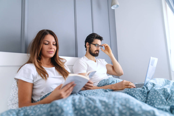 Beautiful Caucasian brunette reading book in bed while her husband with eyeglasses using laptop. Selective focus on man. - Fotó, kép