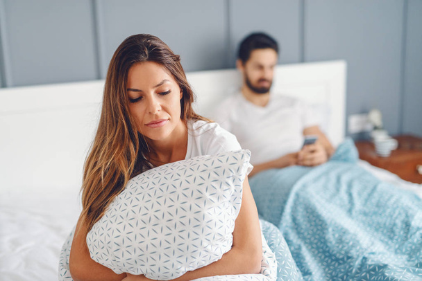 Žárlivý nespokojeni přítelkyně drží polštář a sedí na posteli, zatímco její manžel pro čtení nebo zápis zprávy pomocí chytrého telefonu. Selektivní fokus na ženu. - Fotografie, Obrázek