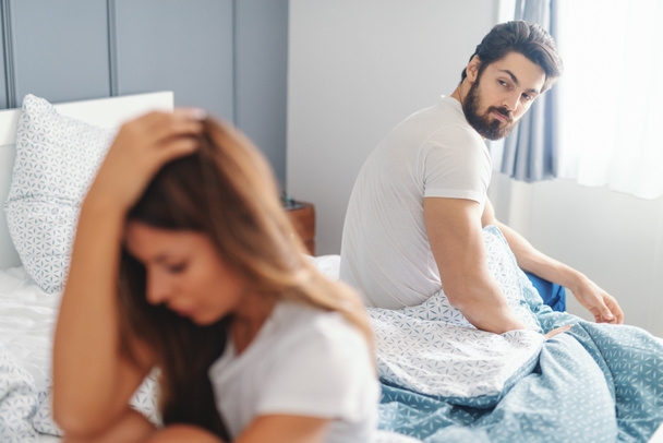 Sad woman sitting on the bed in bedroom in the morning and holding head after fight with husband. Husband looking over shoulder at woman. Selective focus on man. - Photo, Image