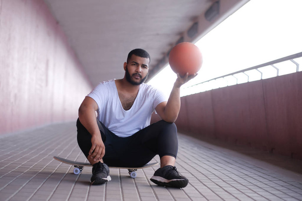 Afro-amerikai ember ül a gördeszka, és tartja a kosárlabda, a híd járdán - Fotó, kép