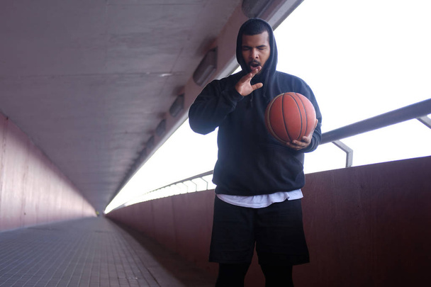Stylish African-American guy wearing a black hoodie holding a basketball and yawning - Photo, image