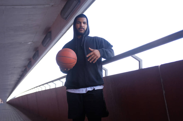 Afrikanisch-amerikanischer Typ mit schwarzem Kapuzenpullover, der einen Basketball hält, während er auf einem Brückensteg steht  - Foto, Bild