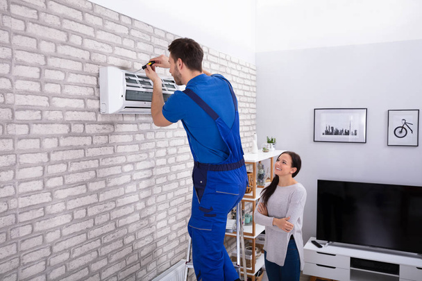 Smiling Woman Looking At Male Technician Repairing Air Conditioner With Screwdriver In Home - Фото, изображение