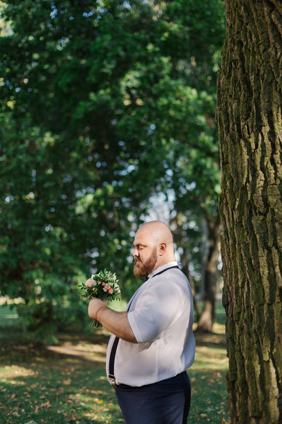 Stylish bearded fat groom with wedding bouquet in the park. - Фото, зображення