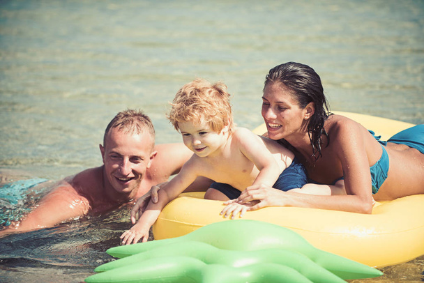 Father and mother near mattress swim with son. Family spend time together and having fun. Cute child boy sits on air mattress pineapple shaped in the ocean with parents. Family vacation concept - Foto, Bild