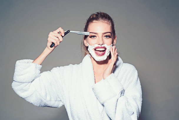 Girl on smiling face wears bathrobe, grey background. Lady play with sharp blade of straight razor. Barber and shaving concept. Woman with face covered with foam holds straight razor in hand - Foto, afbeelding