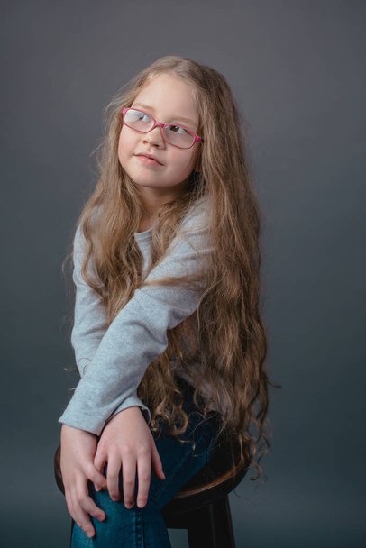 pequena menina bonito adorável com longos cabelos loiros em óculos e em roupas casuais sentado na cadeira no fundo cinza
 - Foto, Imagem