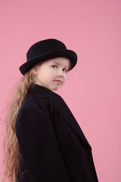 funny cute serious little girl with long blonde hair in black blazer and stylish black hat on pink background - Photo, Image