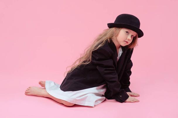 grappige schattig ernstige klein meisje met lang blond haar in zwarte blazer en stijlvolle zwarte hoed op roze achtergrond - Foto, afbeelding