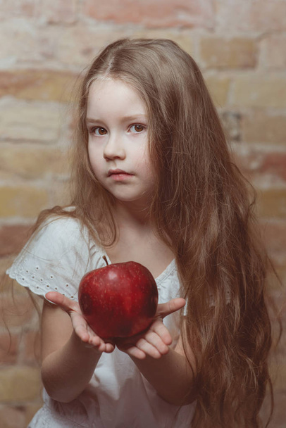 σοβαρό μικρό κορίτσι σε λευκό φόρεμα με κόκκινο μεγάλο μήλο σε τούβλα τοίχο φόντο - Φωτογραφία, εικόνα