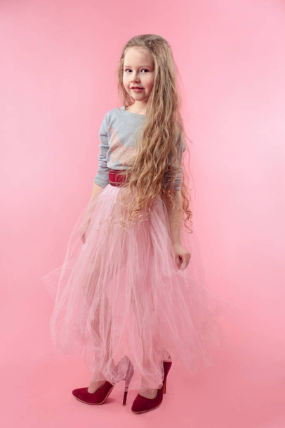 cute adorable little girl with long blonde hair in pink skirt with mom's red high heels shoes playing adult on pink background - Photo, Image