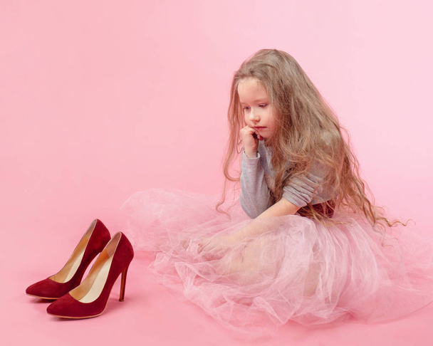 cute adorable little girl with long blonde hair in pink skirt with mom's red high heels shoes playing adult on pink background - Photo, Image