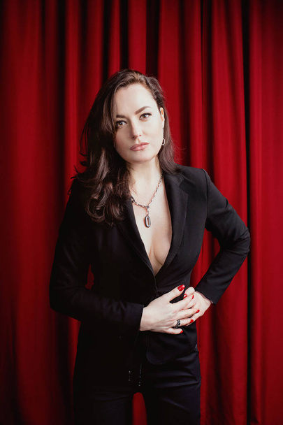 赤いカーテンの背景に黒のスーツのファッショナブルなアクセサリーと美しい豪華なスタイリッシュなブルネットの女性の肖像画 - 写真・画像