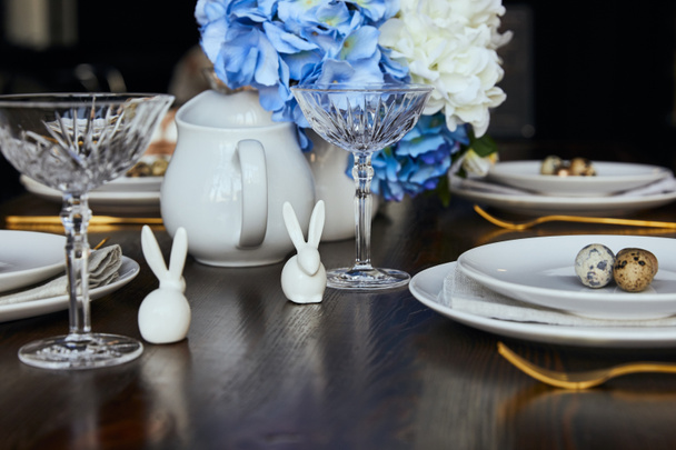 selective focus of decorative rabbits near plates, crystal glass and flowers on wooden table at home - Photo, Image