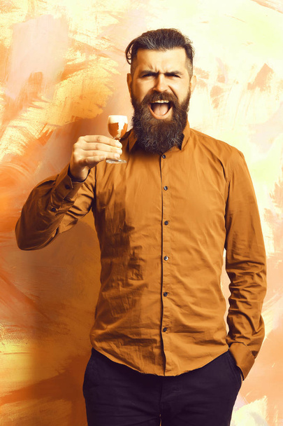 Bearded man, long beard. Brutal caucasian smiling happy hipster with moustache in brown shirt holding alcoholic red shot on colorful texture backgroun - Photo, image