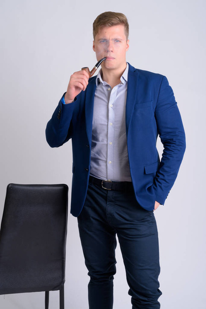 Young handsome blonde businessman smoking pipe while holding chair - Foto, Imagen