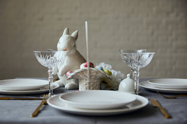 witte borden, bestek, kristallen glazen, decoratieve bunnie en mand met beschilderde eieren op tafel thuis - Foto, afbeelding