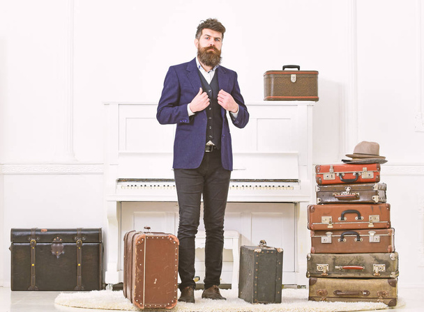 Man, traveller with beard and mustache with luggage, luxury white interior background. Macho elegant on strict face stands near pile of vintage suitcase, ready for vacation. Luggage and travel concep - Foto, Imagem