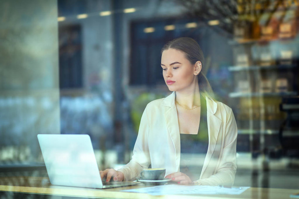 Επιχειρήσεων γυναίκα που εργάζεται στο lap-top στο café, νεαρή γυναίκα κάθεται στο παράθυρο πίνακα στο κυλικείο πίνοντας καφέ και να εργάζονται στον υπολογιστή με τιμολόγιο στο τραπέζι πληρωμή λογαριασμών μέσω του internet banking, παράθυρο αντανάκλαση - Φωτογραφία, εικόνα