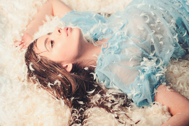 Lady in transparent blue nightie lay on bed in heap of feathers. Woman with long hair in tender pajama relaxing. Girl on mysterious face lay on bed covered with feathers and fluff. Perfume concept - Foto, afbeelding
