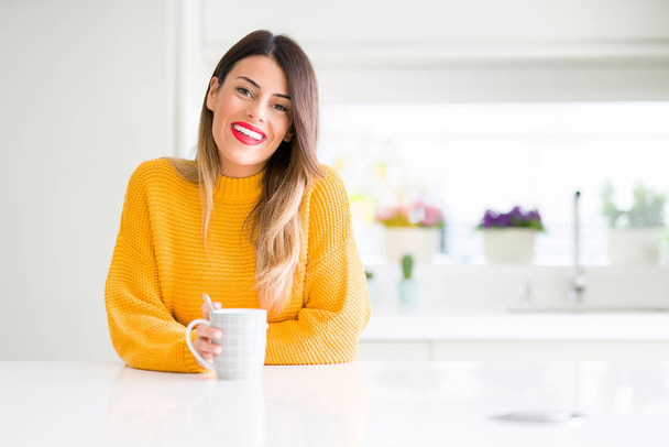 Νεαρή όμορφη γυναίκα πίνοντας ένα φλιτζάνι καφέ στο σπίτι με ένα χαρούμενο πρόσωπο στέκονται και να χαμογελούν με αυτοπεποίθηση χαμόγελο δείχνει τα δόντια - Φωτογραφία, εικόνα